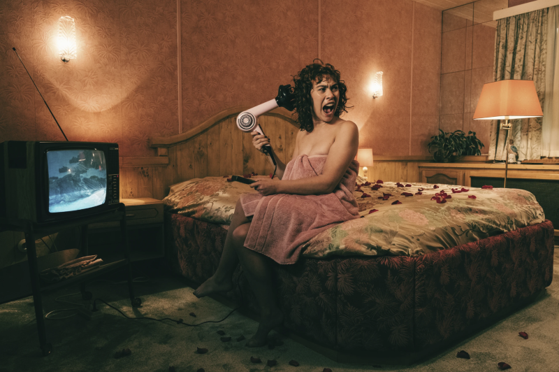 A white woman sits on a motel bed, a hairdryer held to the side of her head, screaming in terror. An old fashioned television broadcasts in the background.